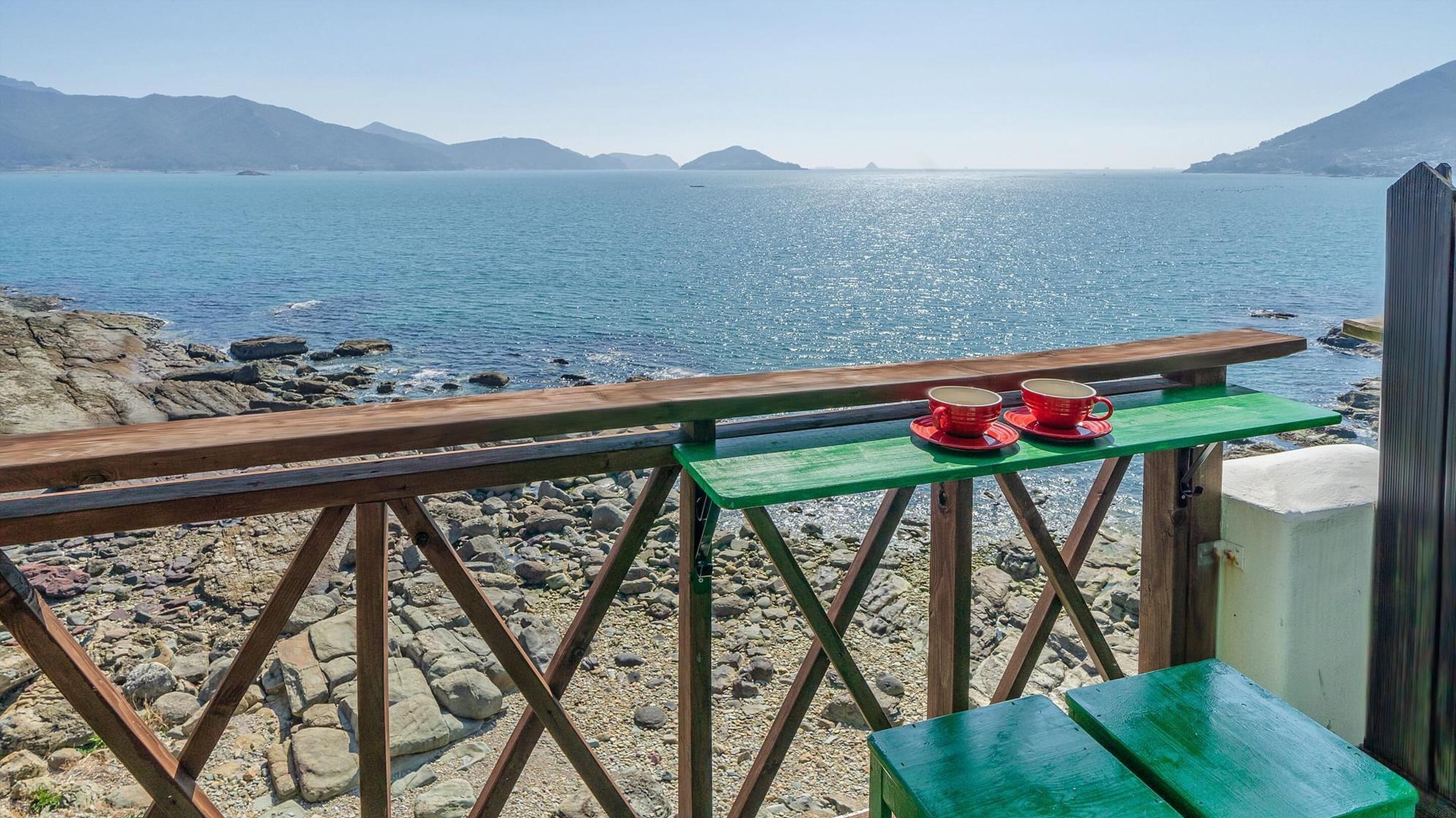 A wooden deck with a green table and two red cups, overlooking a rocky shoreline and calm sea under clear skies.