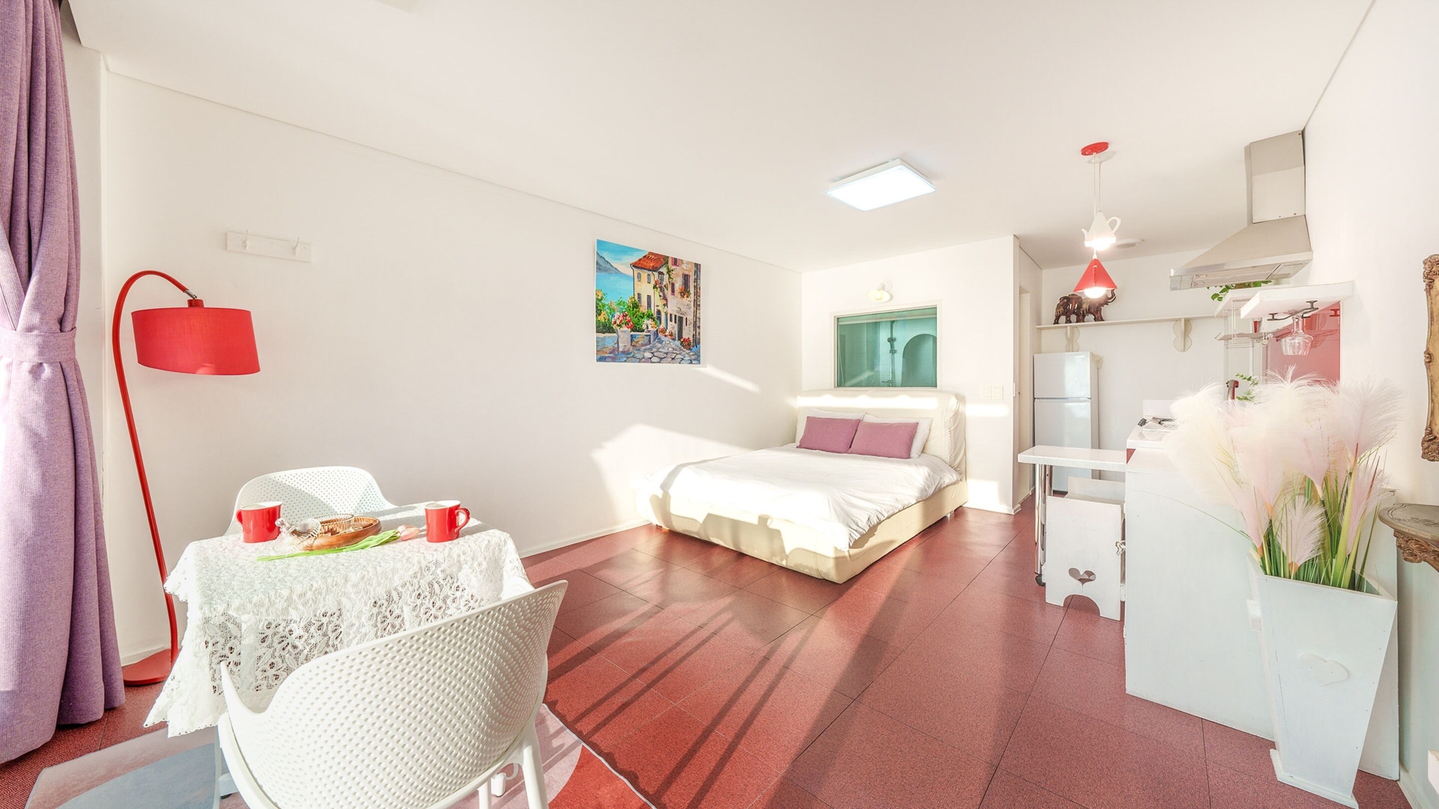 A modern, well-lit room with a bed, a dining table with chairs, a kitchen area, and a painting on the wall.