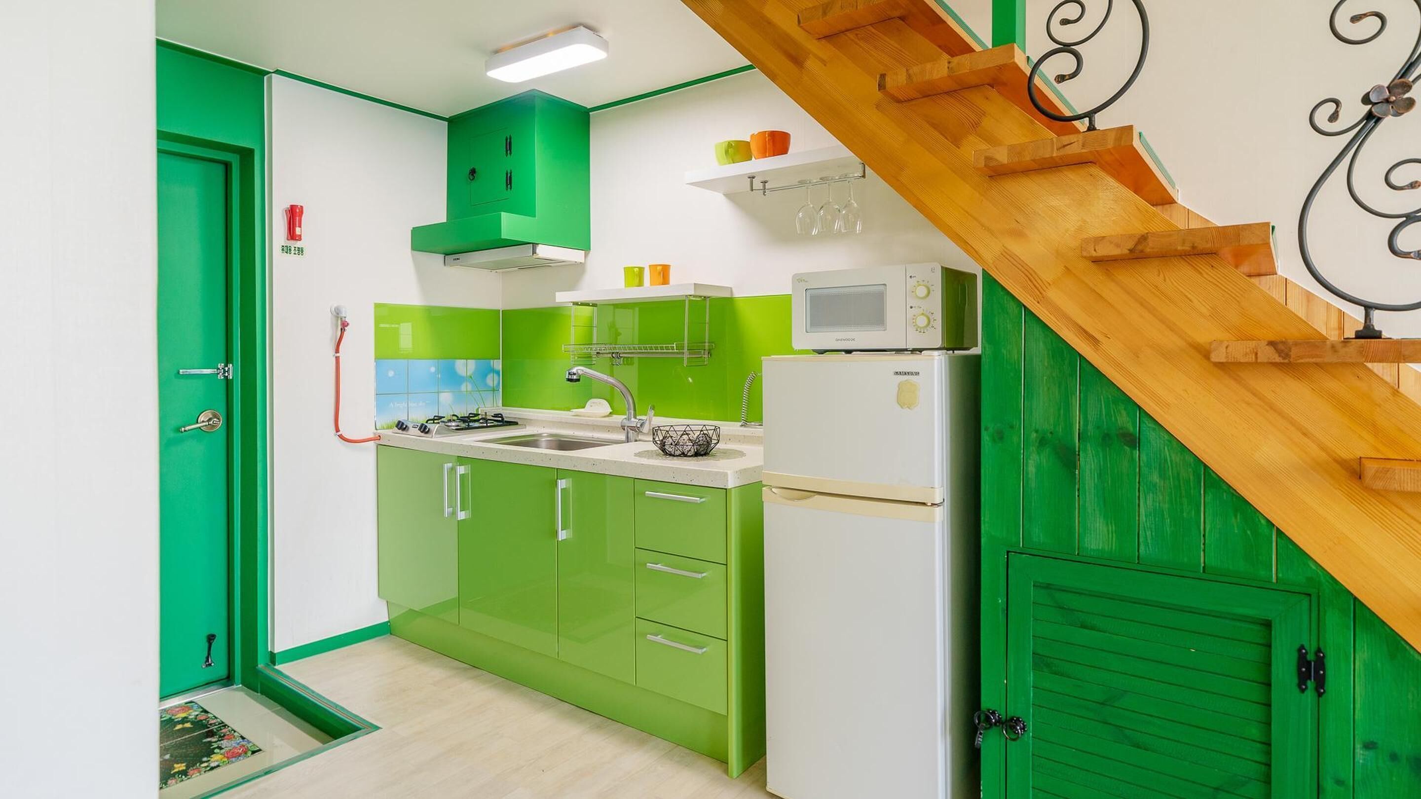 A compact kitchen with green cabinets, a white refrigerator, and a wooden staircase.