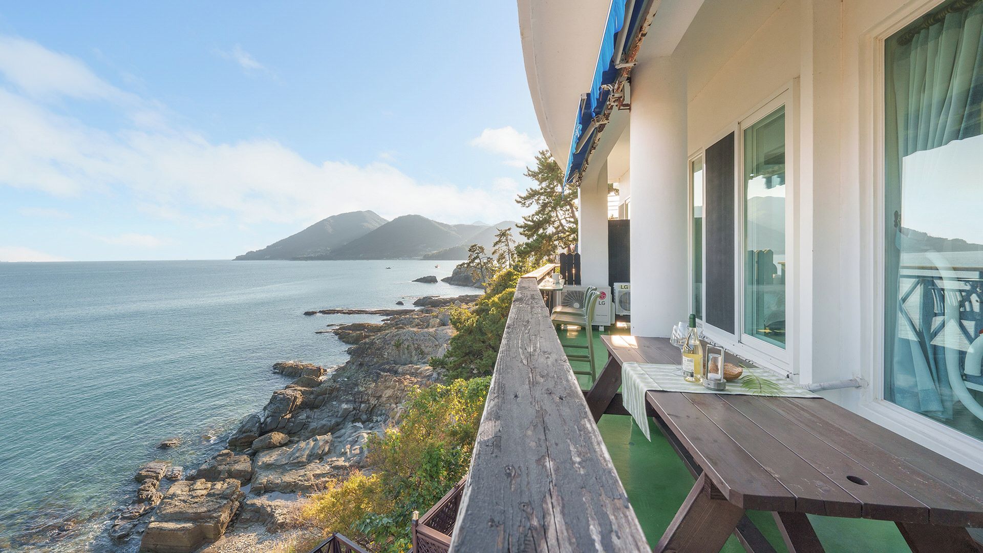 A balcony with a view of the sea and a mountain, wooden furniture, and a table with a bottle and glasses.