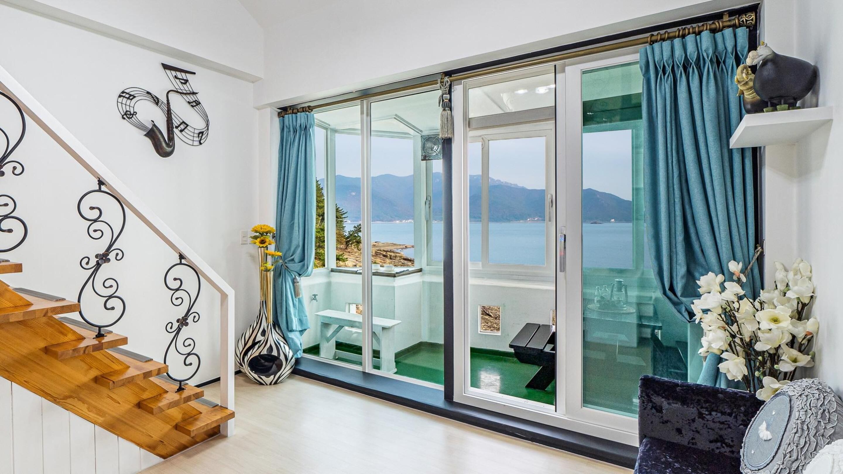 Una sala de estar moderna con una escalera de madera, un ventanal que da a un balcón con vista al mar, y un comedor con mesa y sillas.