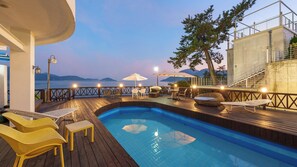 A swimming pool with a wooden deck, outdoor seating, and a view of the sea at dusk. - Namhae Opera House Pension (Namhae)