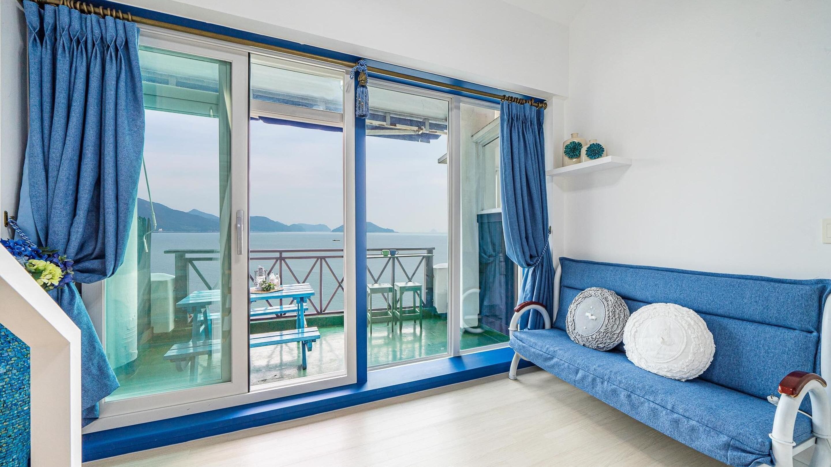 A room with a large sliding glass door leading to a balcony with a table and chairs, a blue sofa with decorative pillows, and a white shelf with decorative items.
