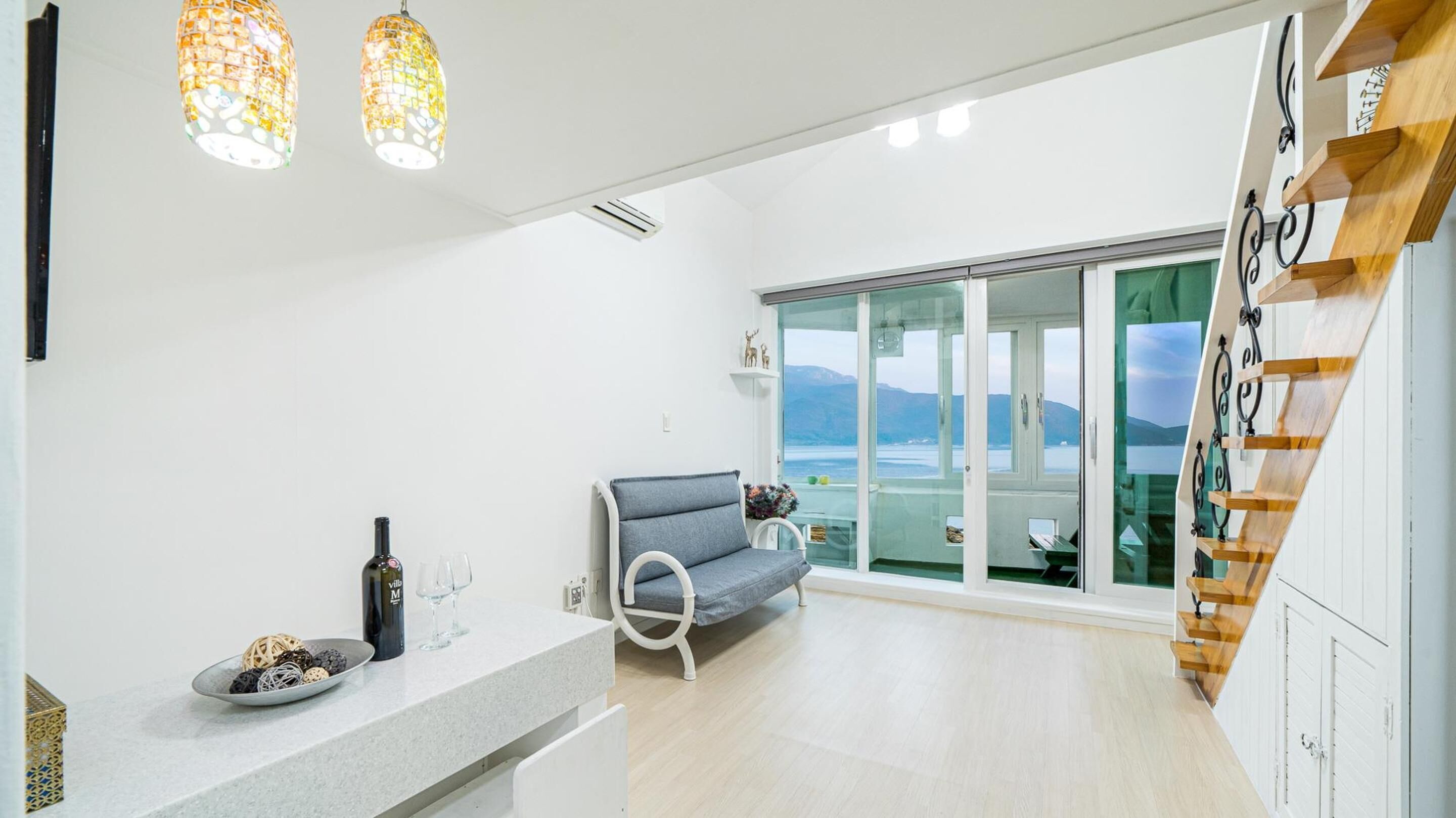 A modern kitchen with a built-in oven, a dining area with a grey sofa, and a view of the sea through large glass doors.