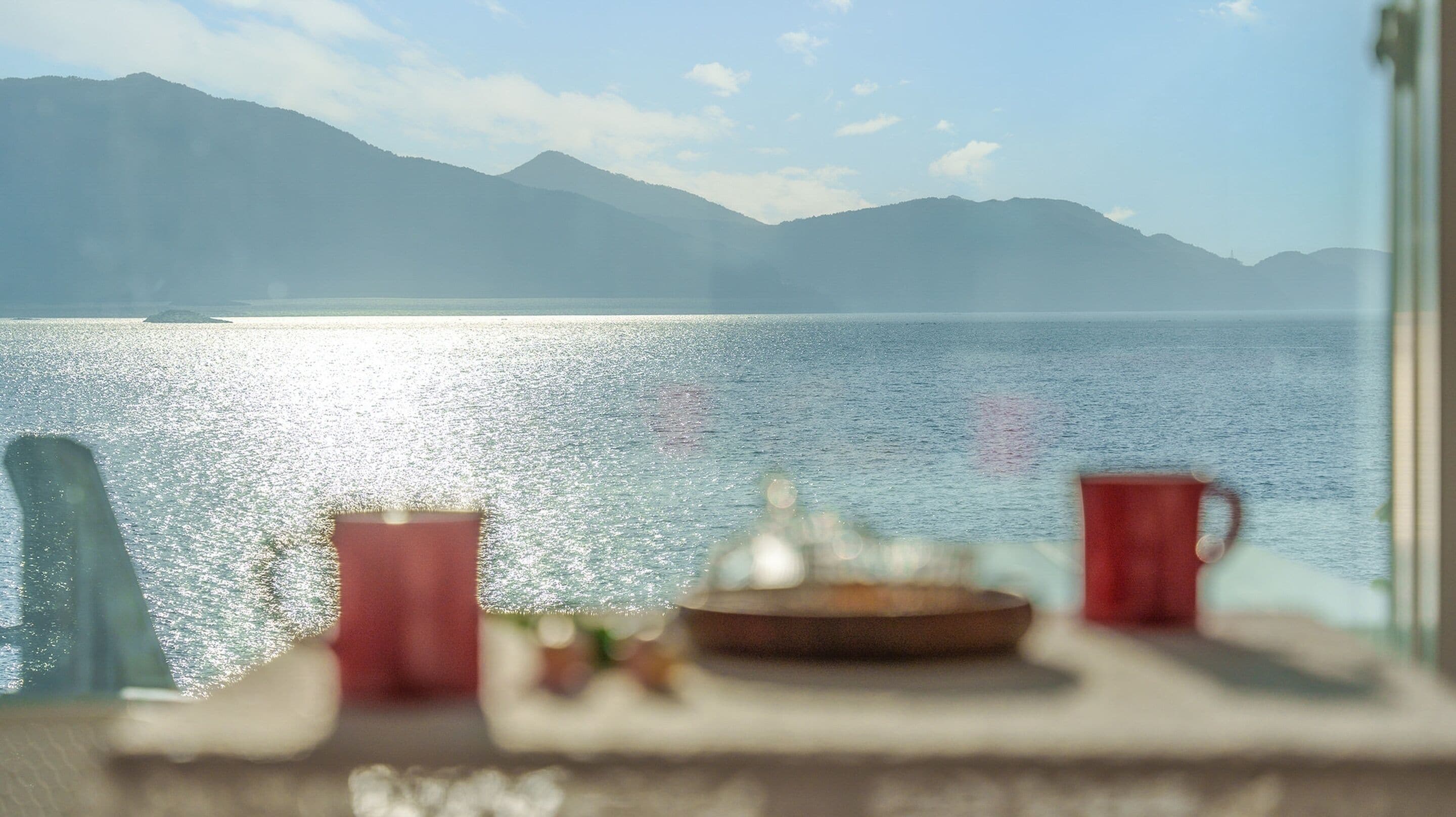A table with a red mug, a glass, and a plate, overlooking a calm lake with mountains in the background.