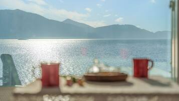 A table with a red mug, a glass, and a plate, overlooking a calm lake with mountains in the background.