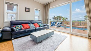 A modern living room with a large window, a sofa with colorful cushions, a coffee table, and a view of a pool and palm trees.