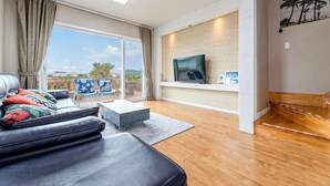 A modern living room with a leather sofa, a coffee table, and a TV on a wall. There is a balcony with a view of the city and palm trees.