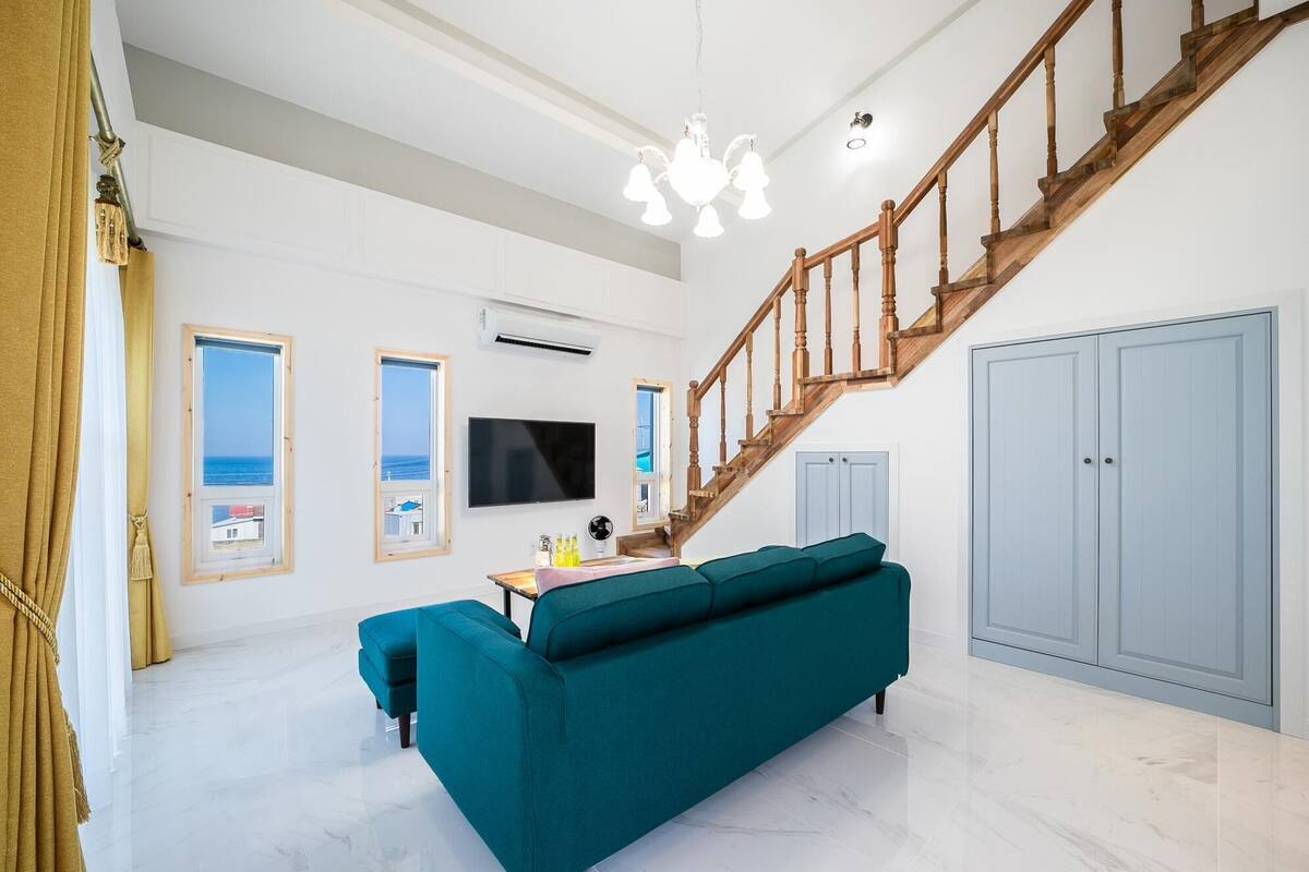 A spacious living room with a teal sofa, a wooden staircase, a flat-screen TV, and a view of the sea through windows.