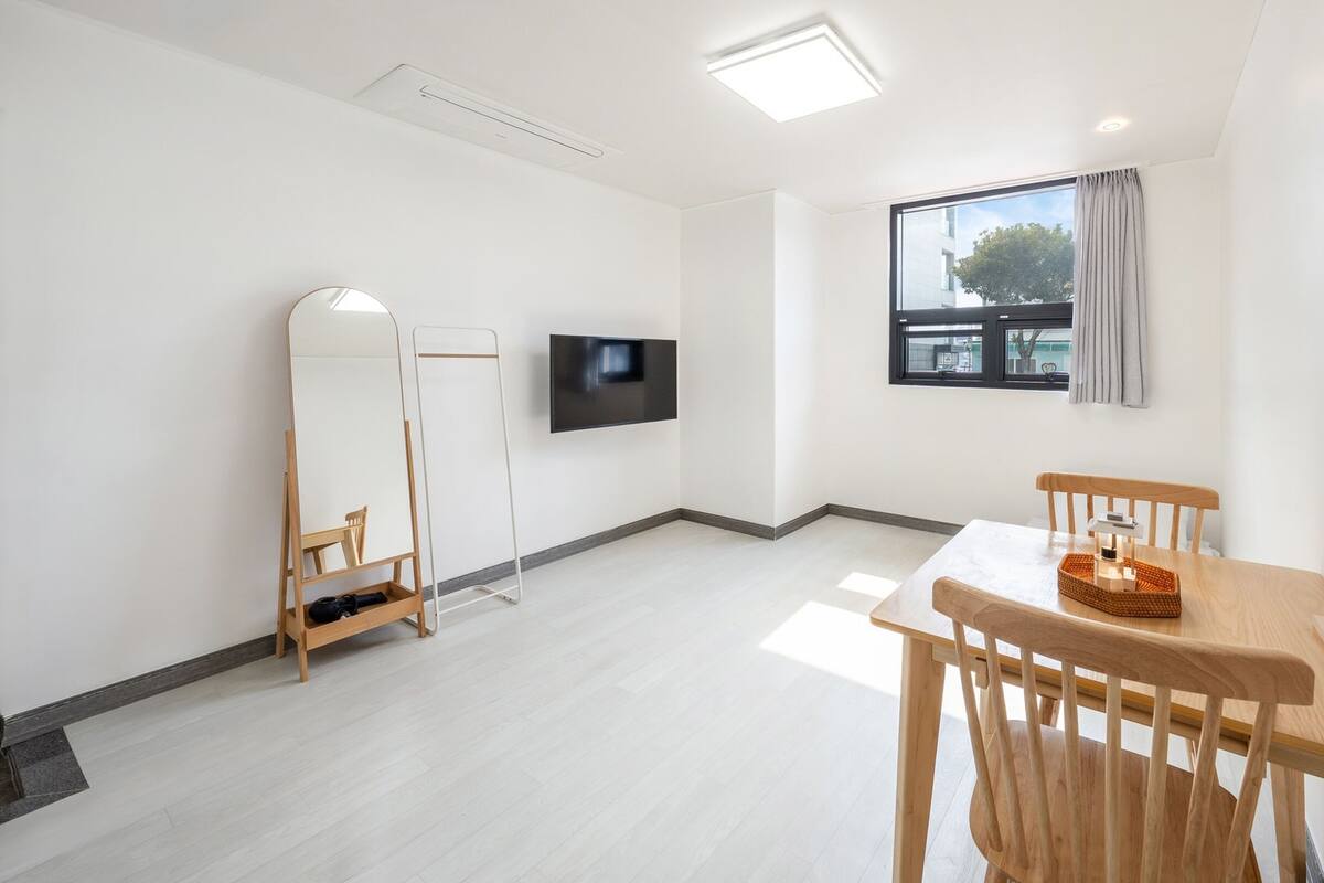 Una habitación moderna y minimalista con una televisión de pantalla plana, una mesa de comedor de madera con sillas, un espejo de pie y una ventana con vistas al exterior.