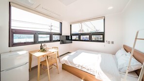 A compact hotel room with a bed, a desk, and a chair. There are two windows providing a view of the cityscape. - Yeosu Nangmanstay Pension (Yeosu)