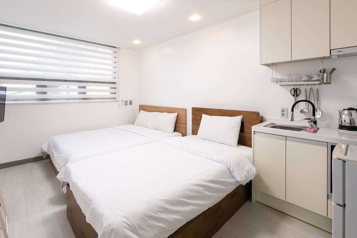Habitación de hotel con cama, televisor, kitchenette y ventana con persianas.