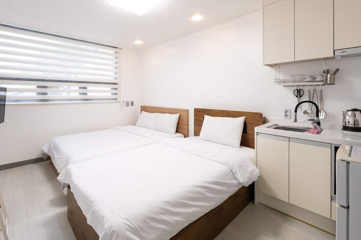 A hotel room with a bed, a TV, a kitchenette, and a window with blinds.
