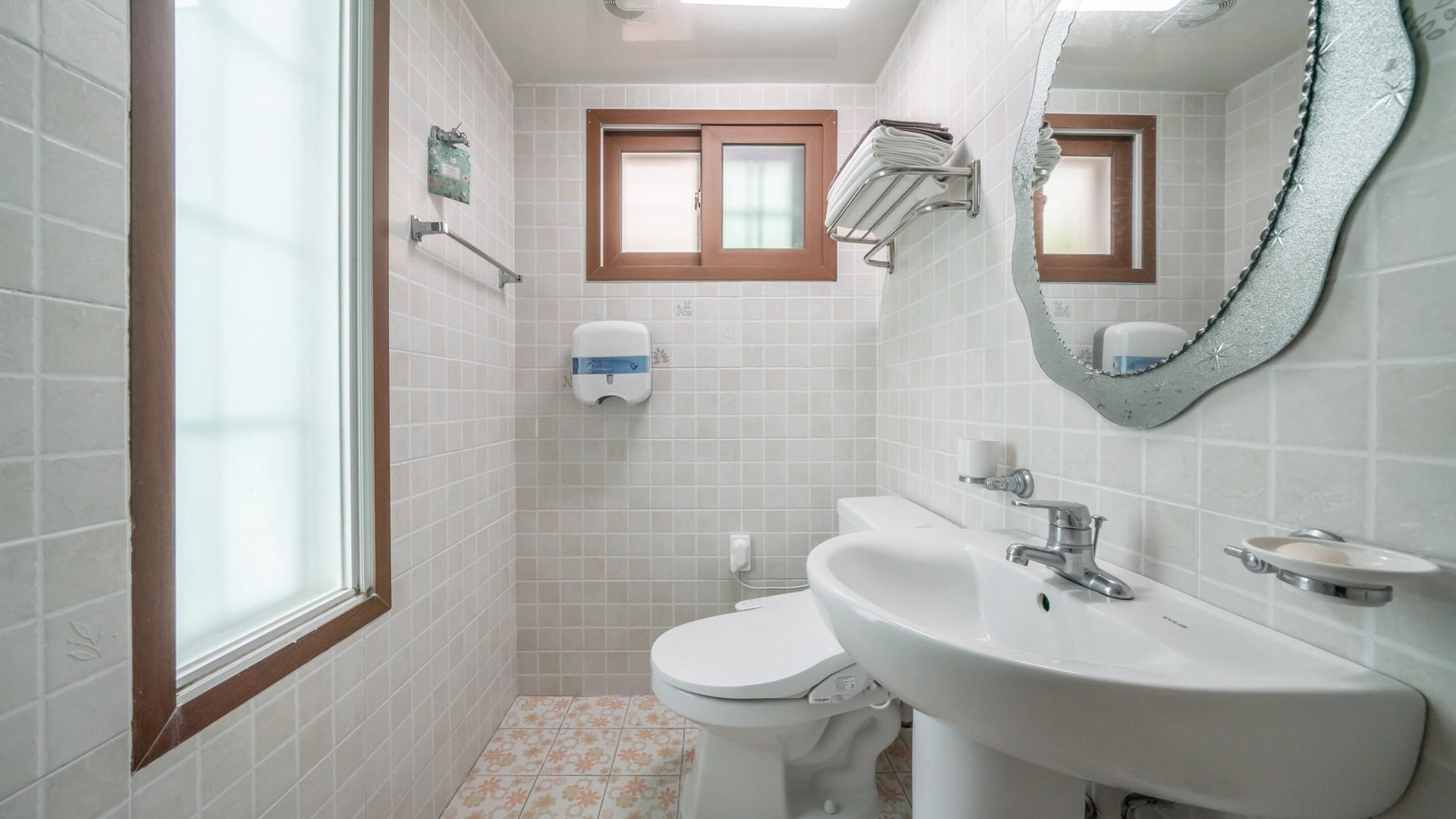 Un baño con azulejos blancos, un inodoro, un lavabo, un espejo, una ventana y un perchero para toallas.