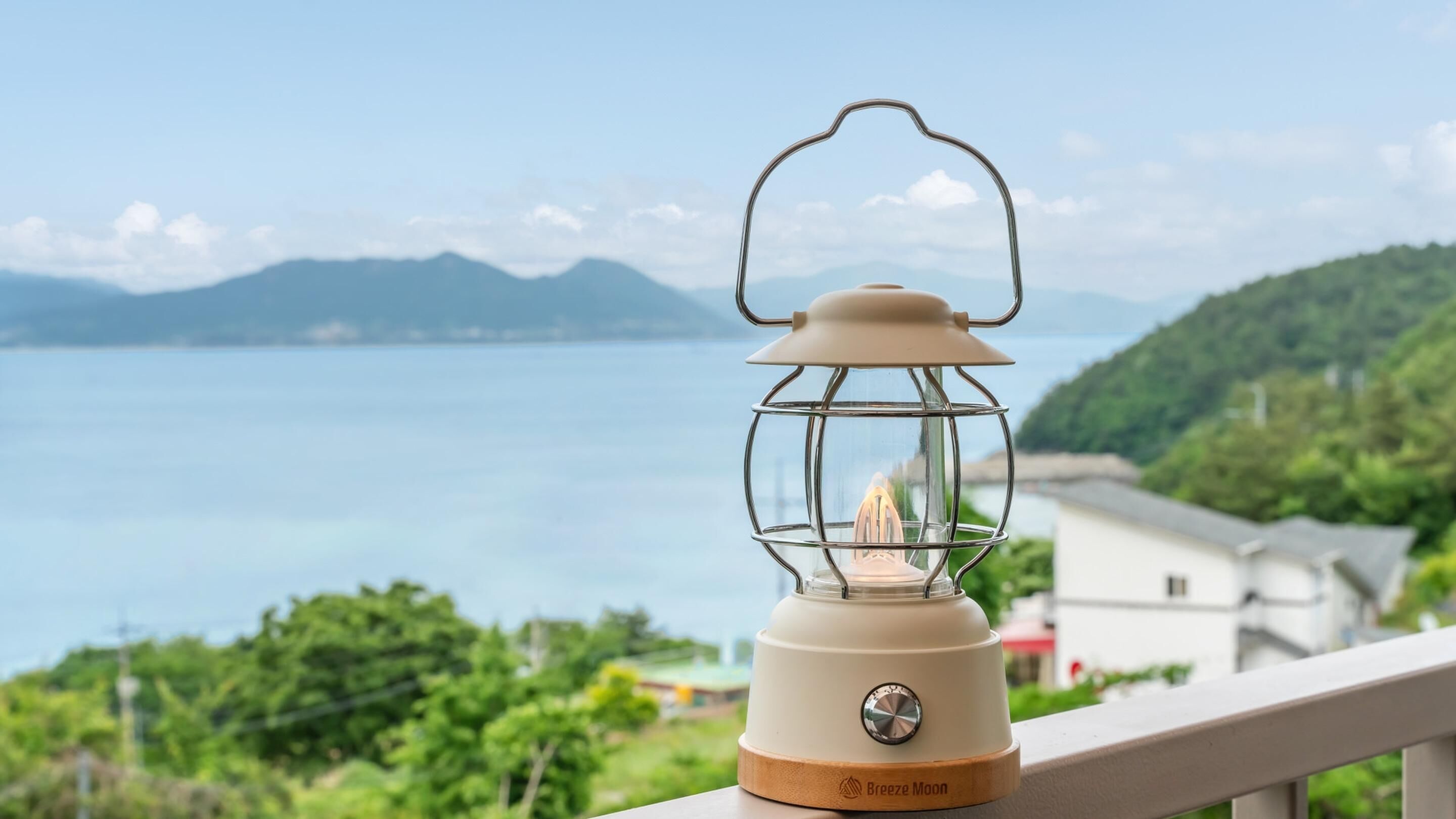 A beige lantern with a metal frame and a wooden base, placed on a balcony overlooking a body of water and greenery.