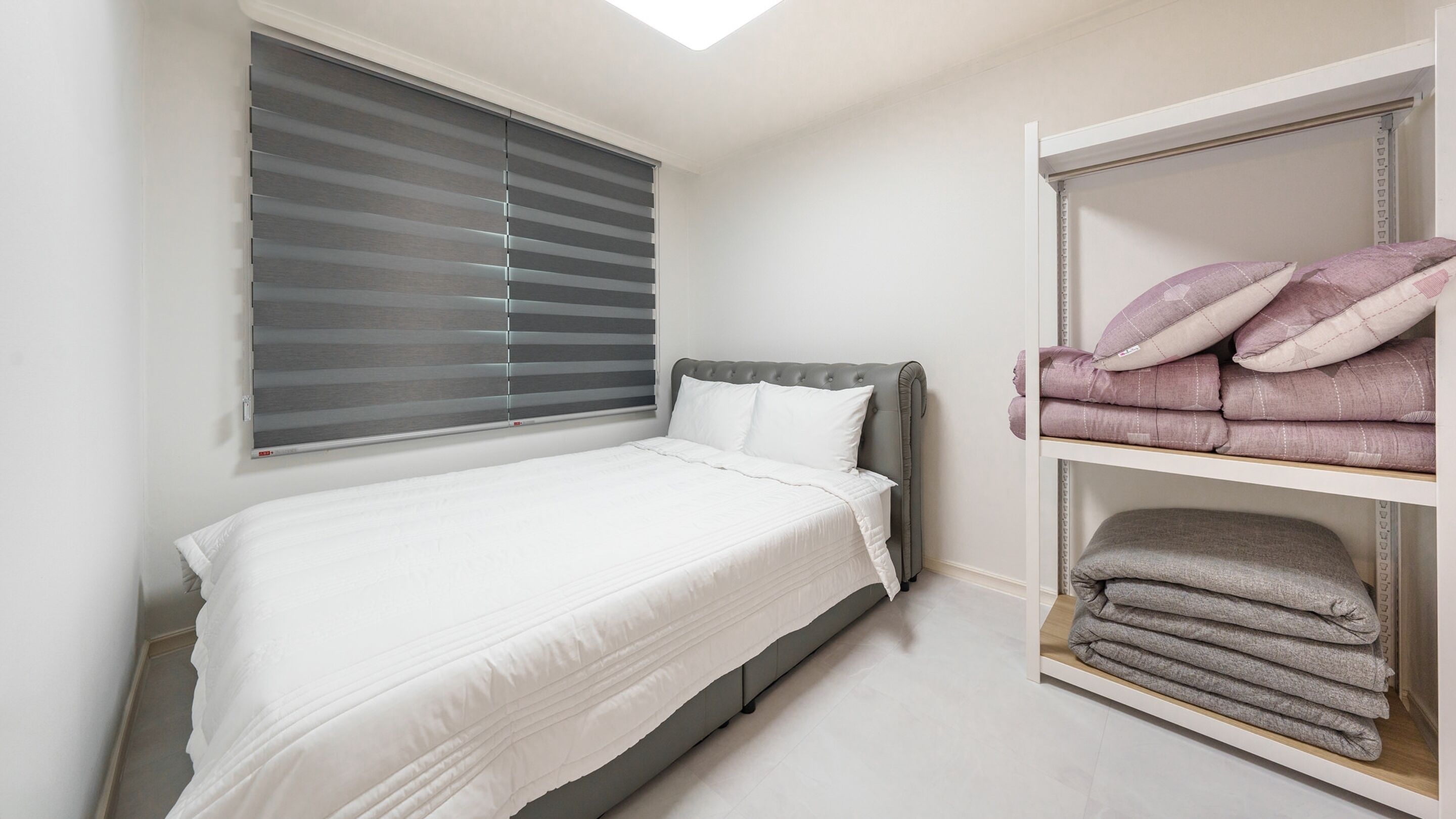 A bedroom with a bed, a window with blinds, and a wardrobe with folded clothes.