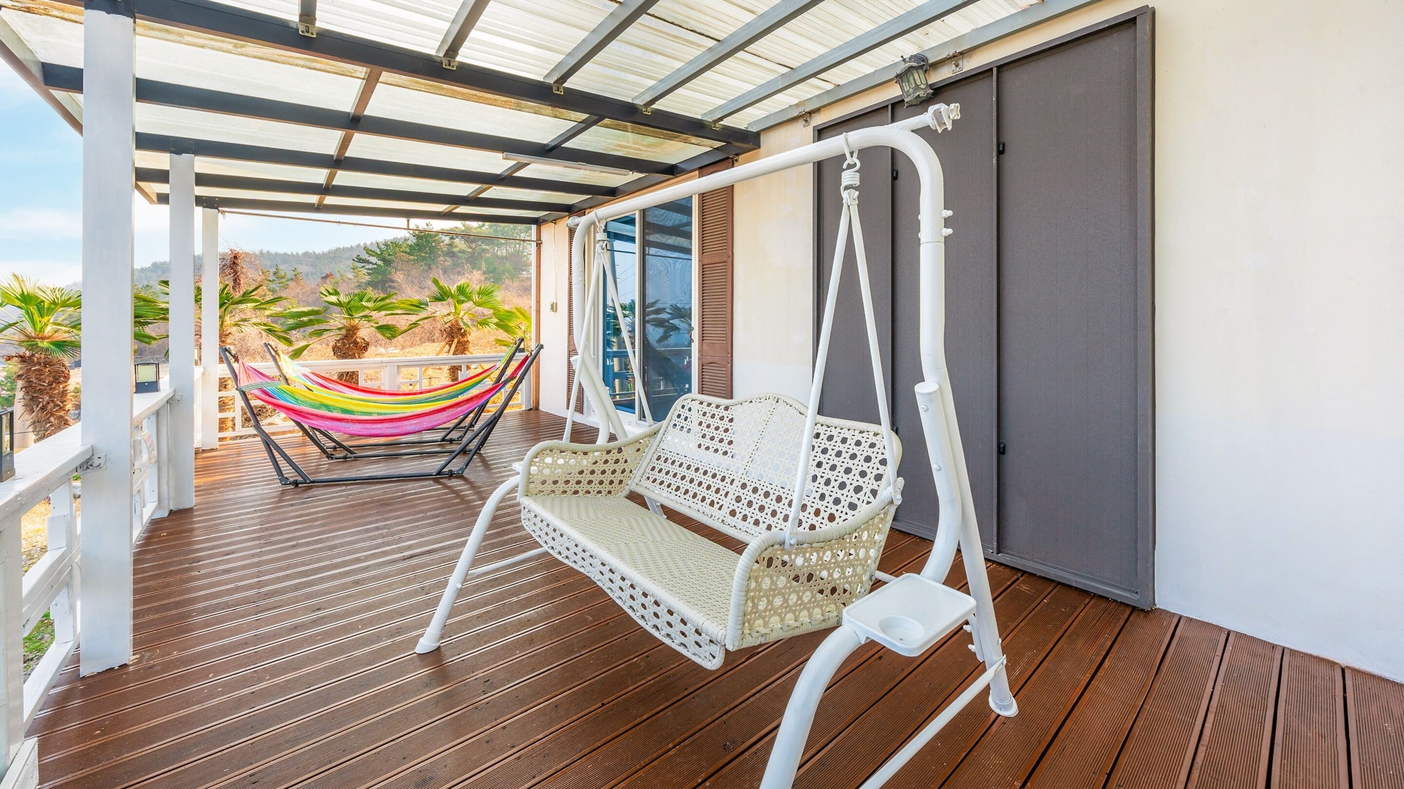 A covered outdoor area with a wooden deck, a white wicker swing chair, a colorful hammock, and a view of palm trees and hills.