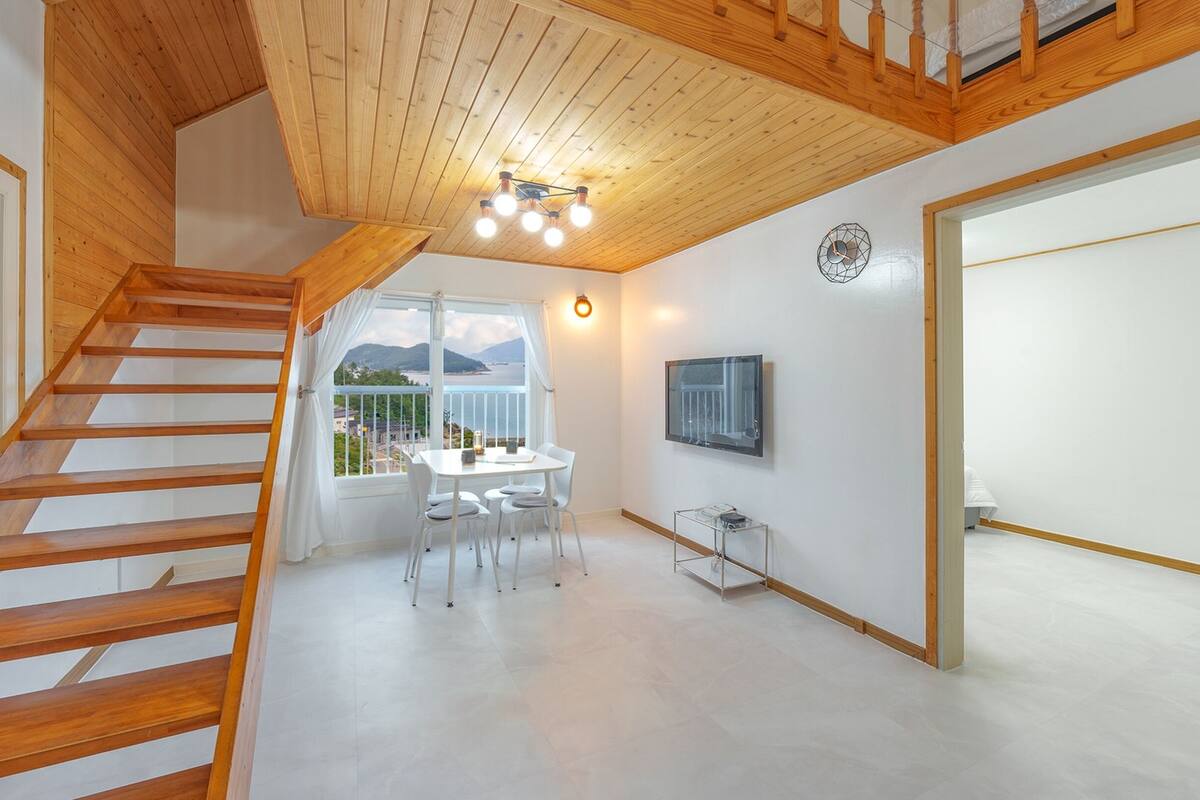 A modern interior with a wooden staircase, a dining area with a table and chairs, a mounted TV, and a view of the outdoors.