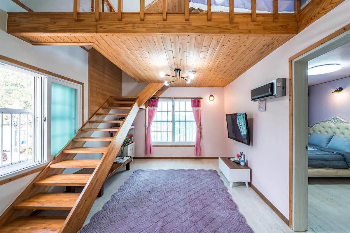 A spacious room with a wooden staircase, a large window, a flat-screen TV, and a cozy bed.