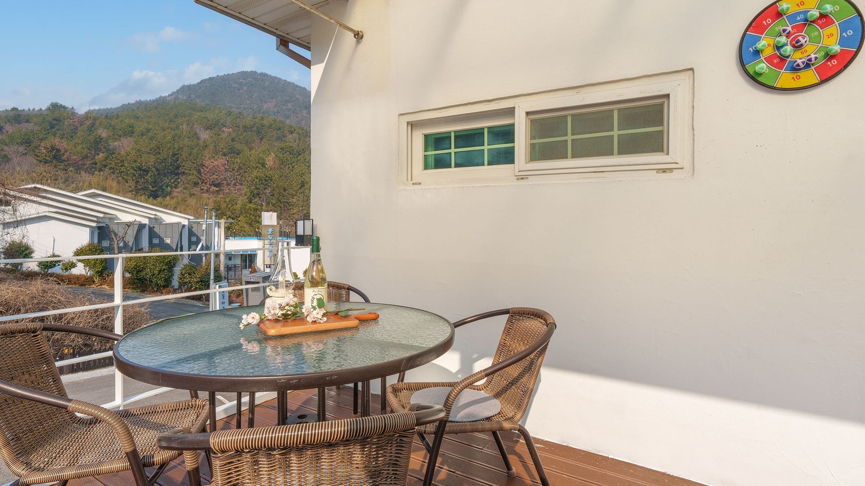 A patio with wicker furniture, a table with a bottle and flowers, a dartboard, and a view of buildings and hills.