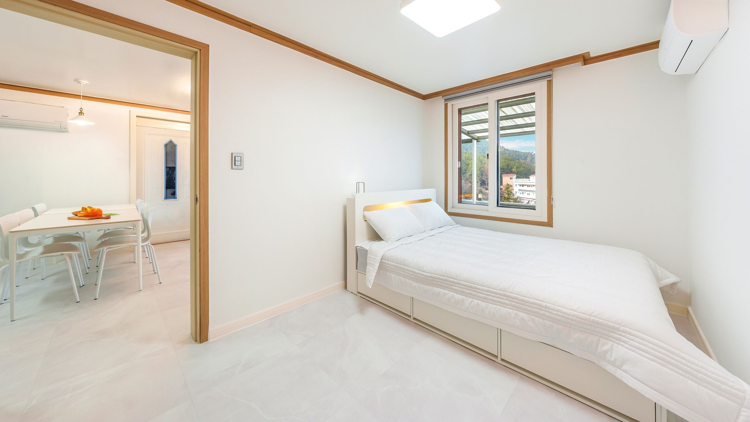 A bedroom with a bed, a window with a view, a desk with chairs, and a door leading to another room.