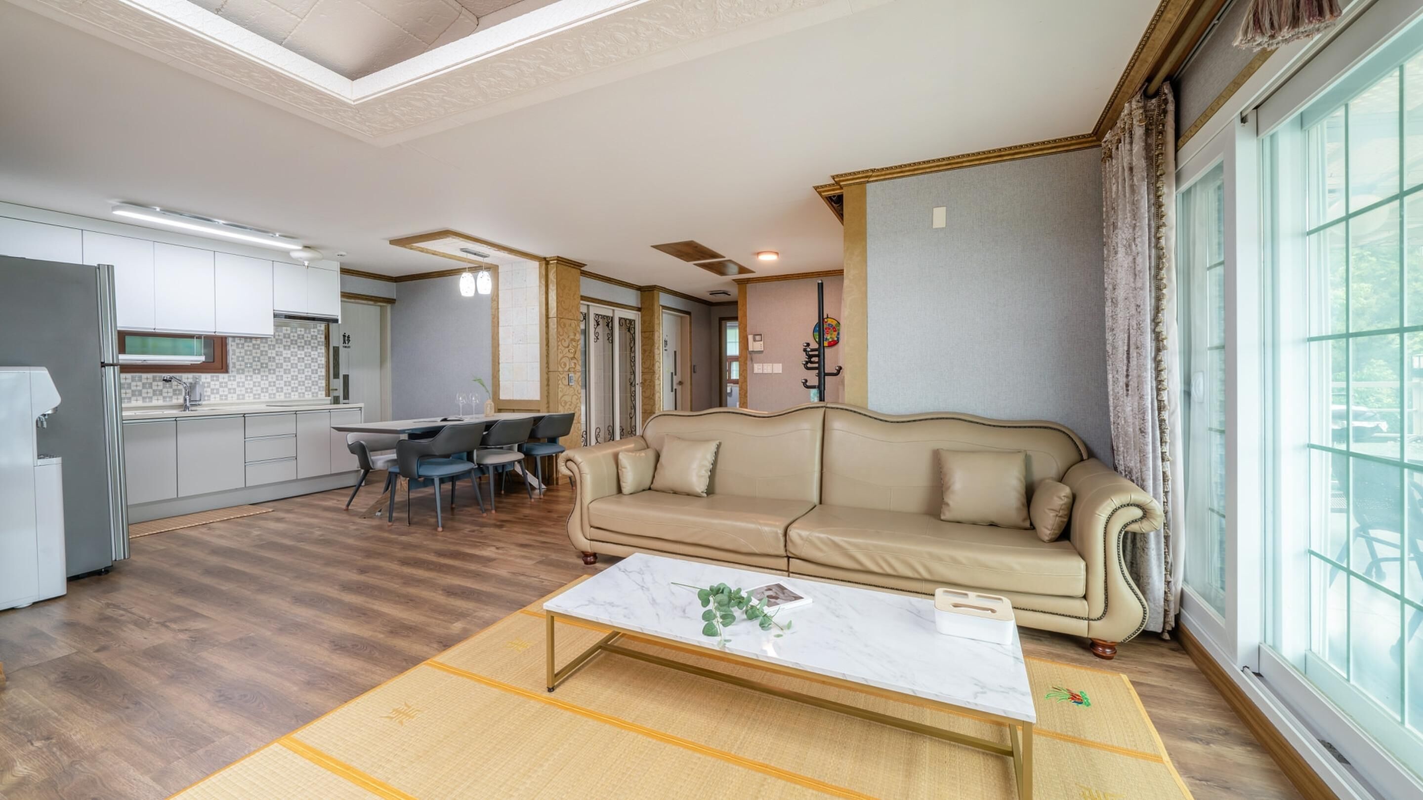 Een moderne woonkamer met een bank, een zithoek met stoelen, een keuken met kastjes, en een groot raam.