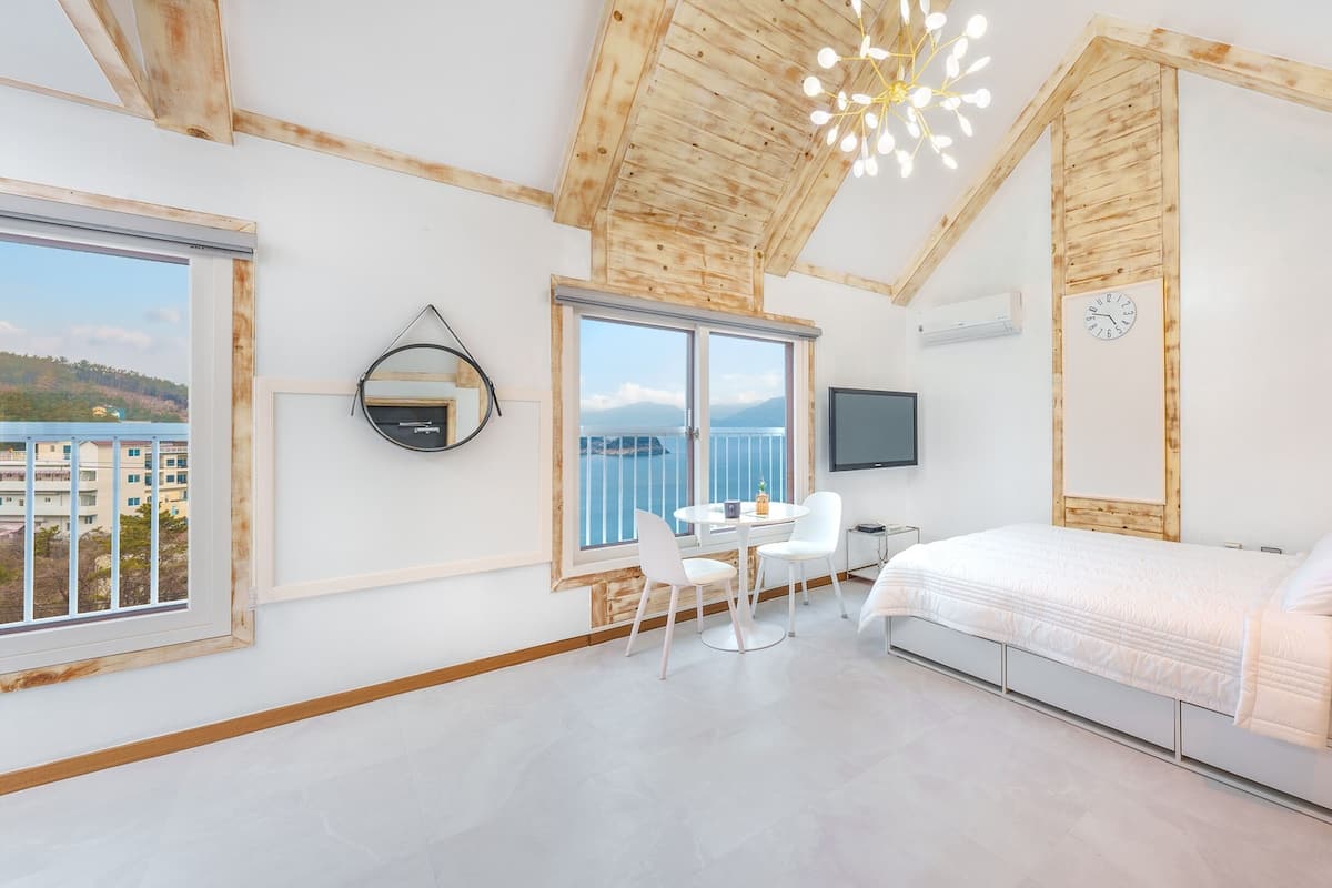 A modern bedroom with a large bed, a TV, a desk with chairs, and a balcony with a view of the sea.