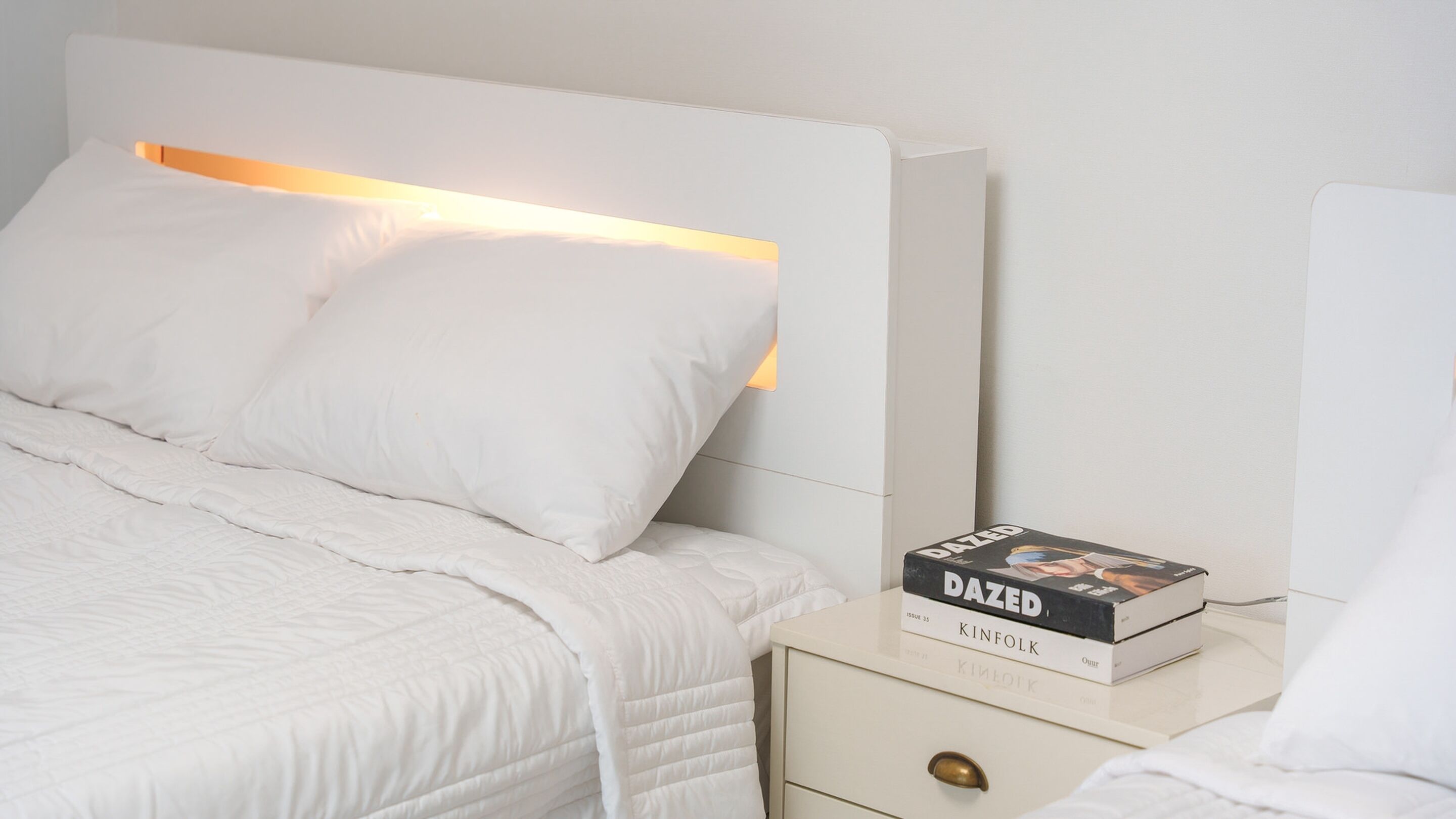 A neatly made bed with white bedding and pillows, a bedside table with a book titled 'DAZED' by Kinfolk, and a nightstand with a drawer.