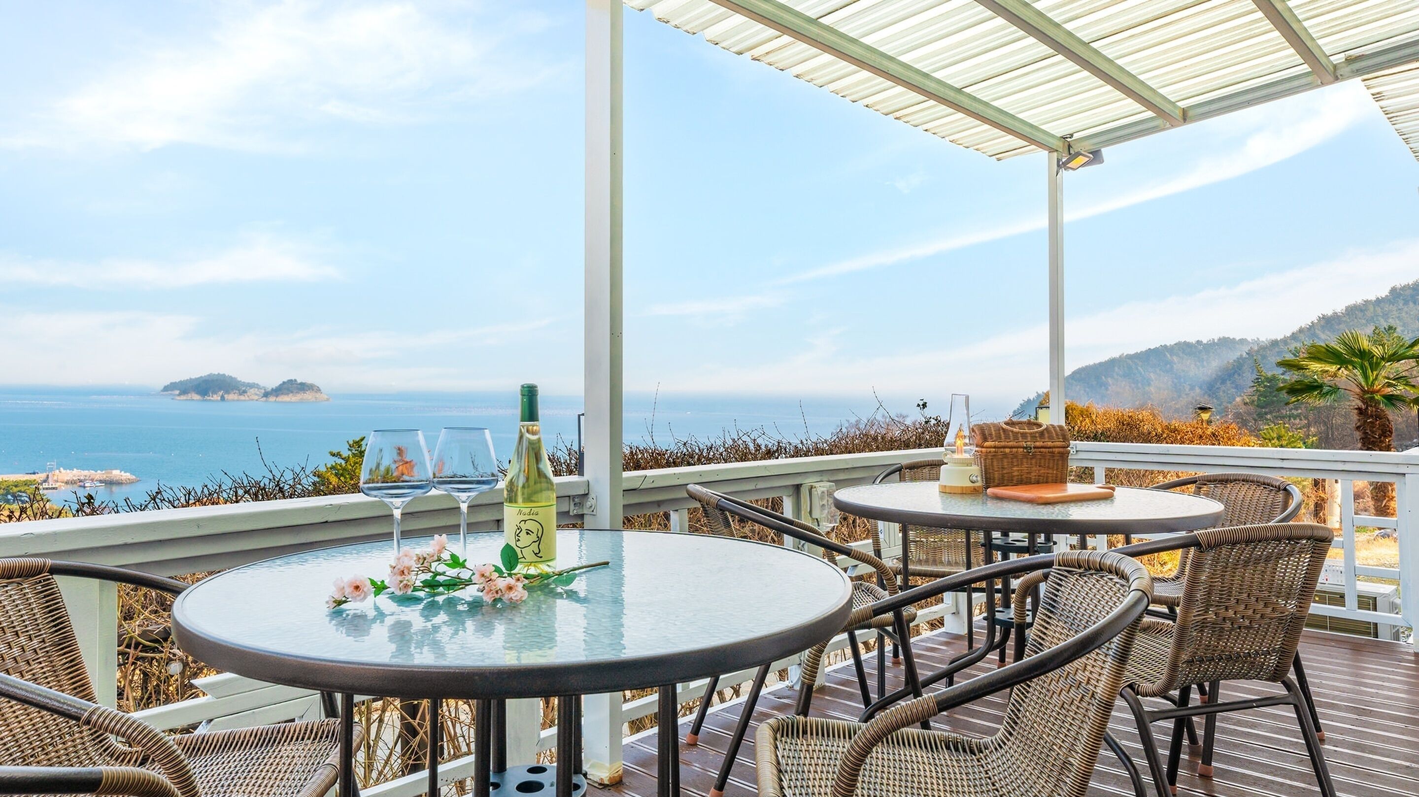 A covered outdoor seating area with two round tables, wicker chairs, a bottle of wine, and a glass on one table.
