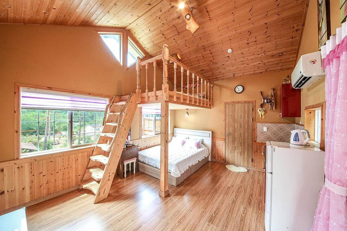 A cozy cabin with a loft bedroom, a wooden staircase, a kitchen area, and a large window offering a view of the outdoors.