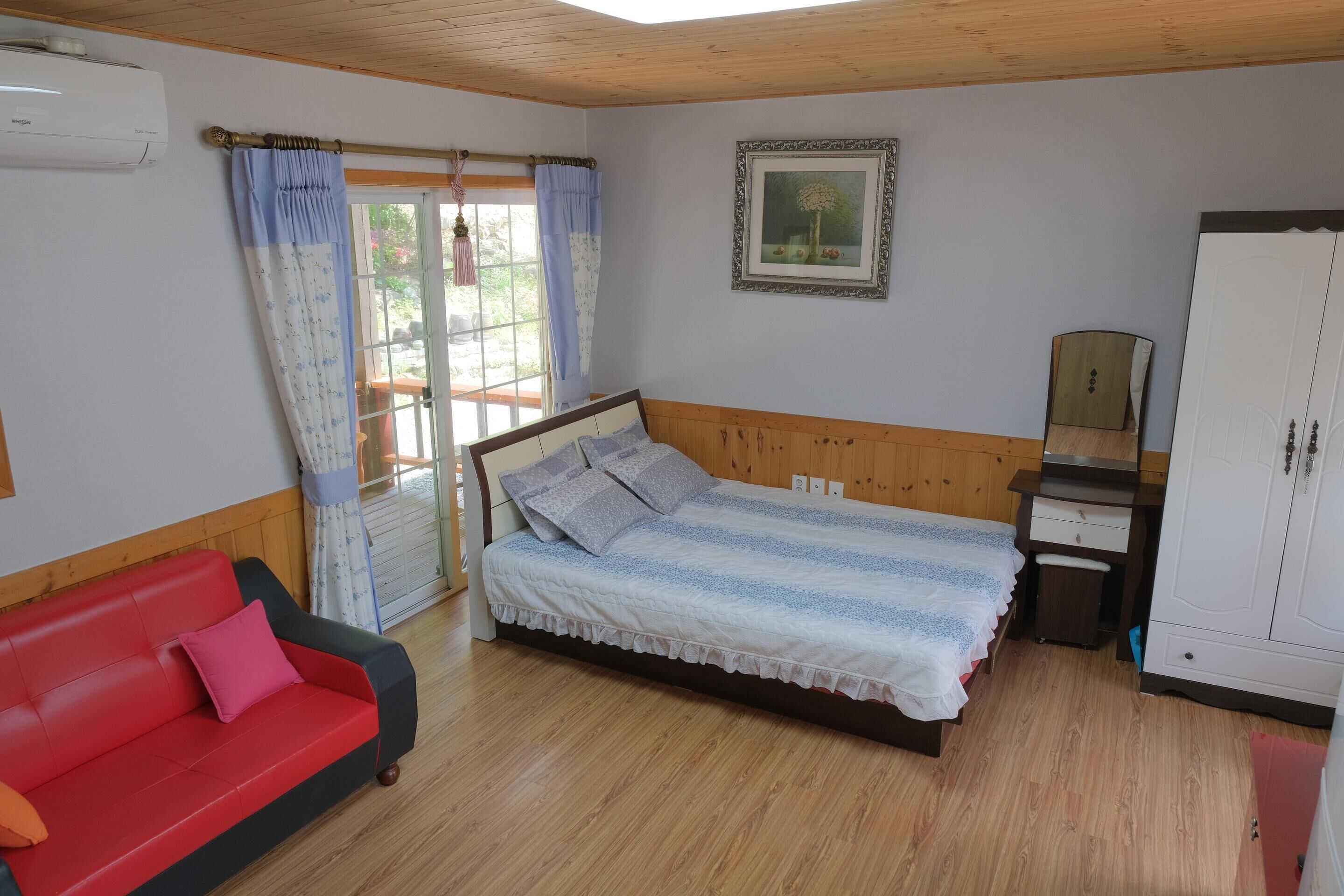 A bedroom with a bed, a red sofa, a wooden wardrobe, and a window with curtains.
