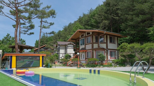Una piscina con un bordo in legno, circondata da una zona boschiva e un edificio a più piani con un balcone.