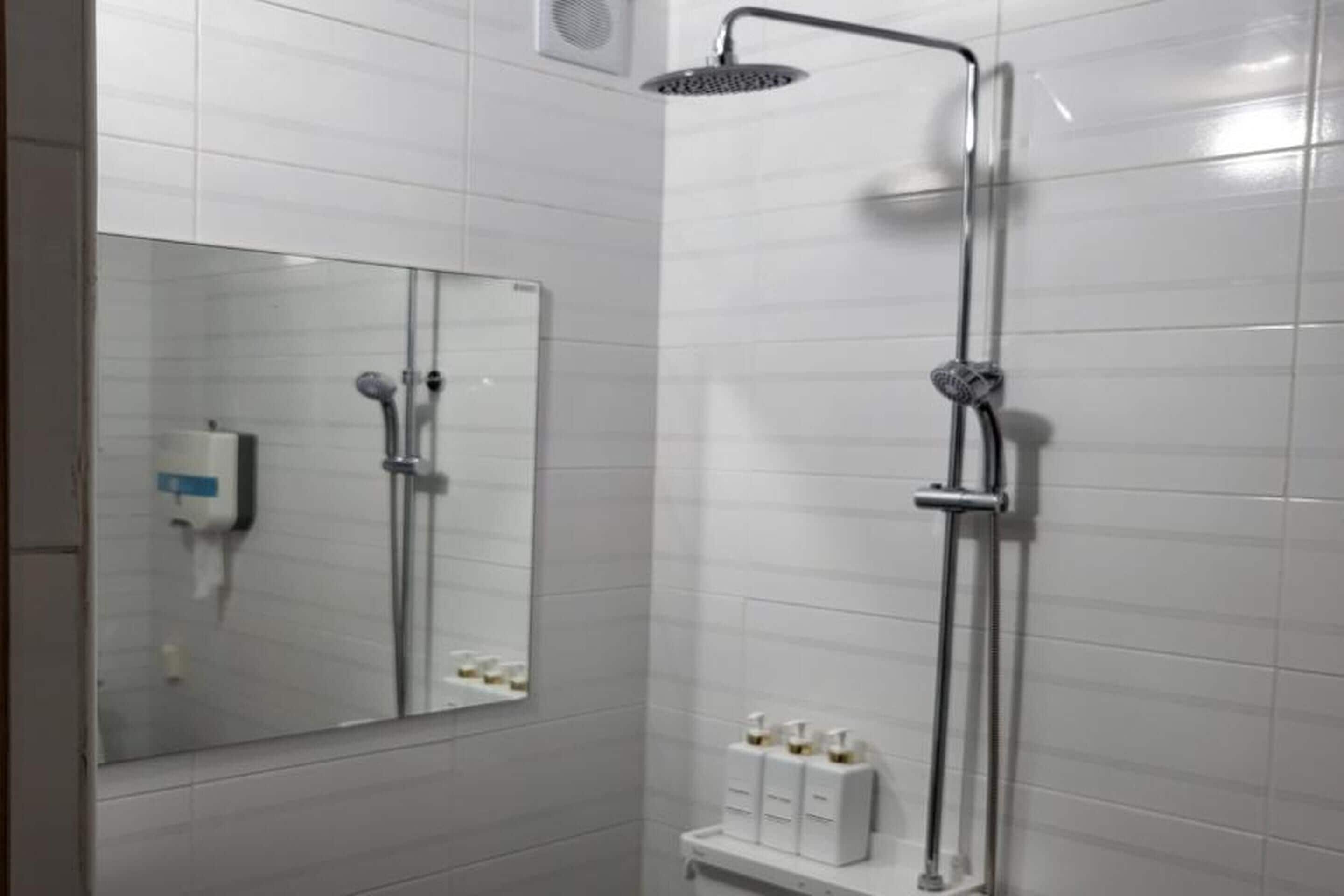 Un baño moderno con azulejos blancos, un espejo grande, una ducha con cabezal de ducha manual y un estante con artículos de tocador.