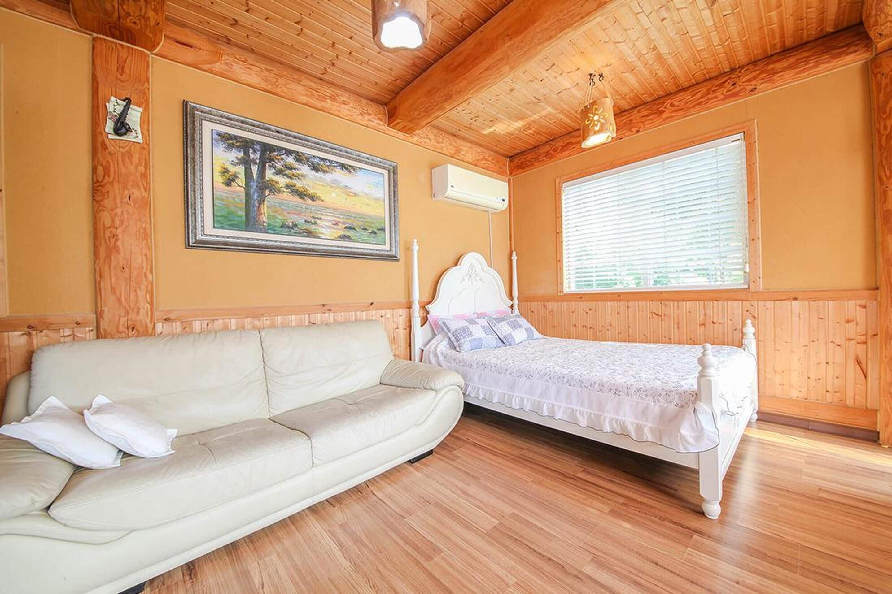 A room with a wooden ceiling, a bed with a white headboard, a beige sofa, and a painting on the wall.
