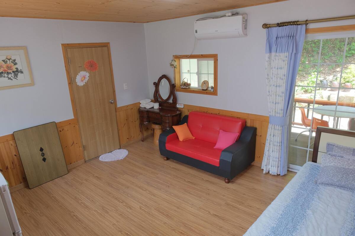 A room with wooden walls and flooring, a red and black sofa, a wooden dresser, a mirror, and a window with a curtain.