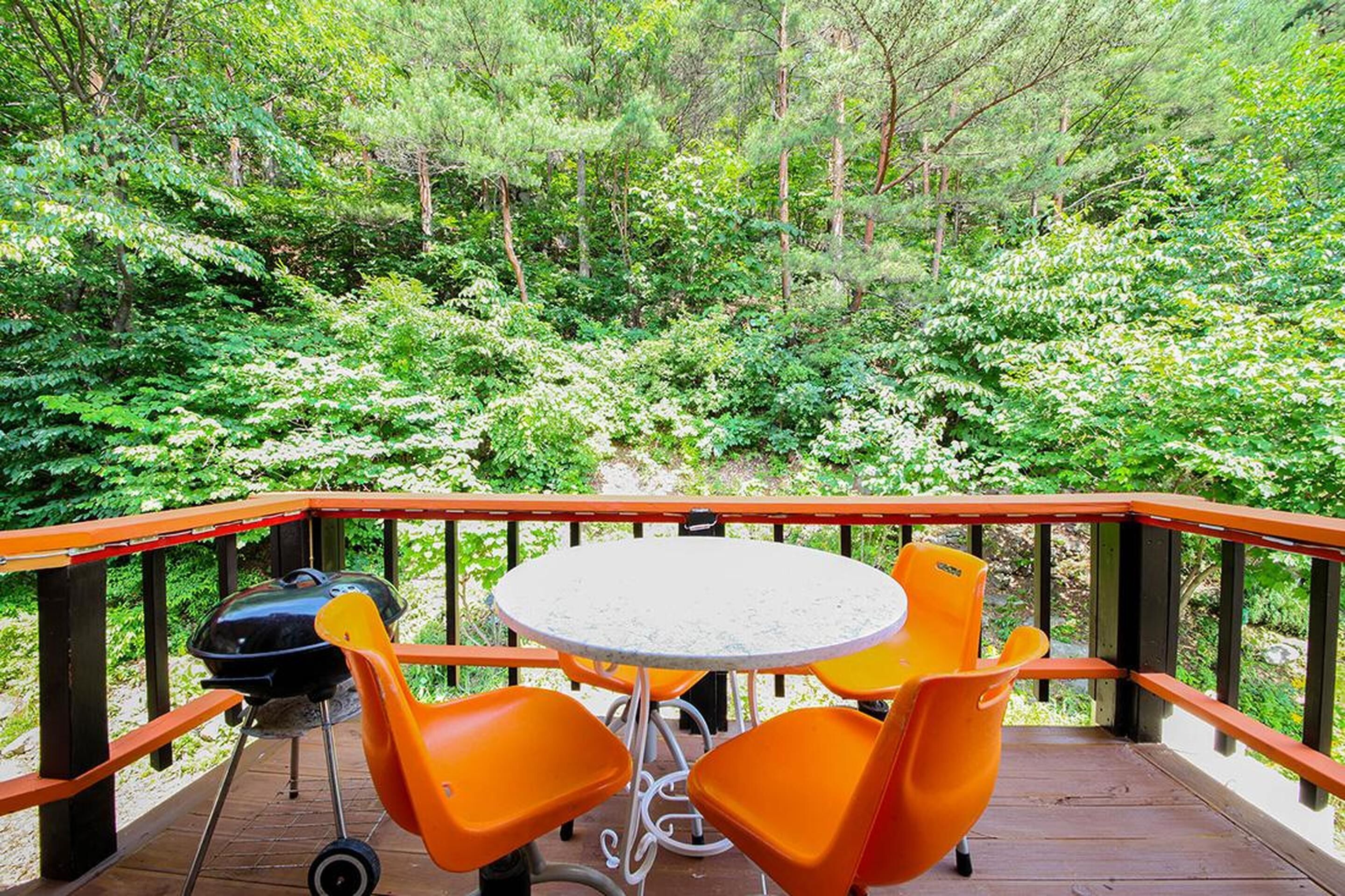 Una terraza con una mesa redonda y sillas anaranjadas, rodeada de una barandilla de madera, con vistas a un bosque verde y frondoso.