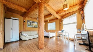 Un'ampia stanza con pavimento in legno, un'imponente colonna di legno, un divano bianco, un letto, una scrivania e una finestra con persiane.
