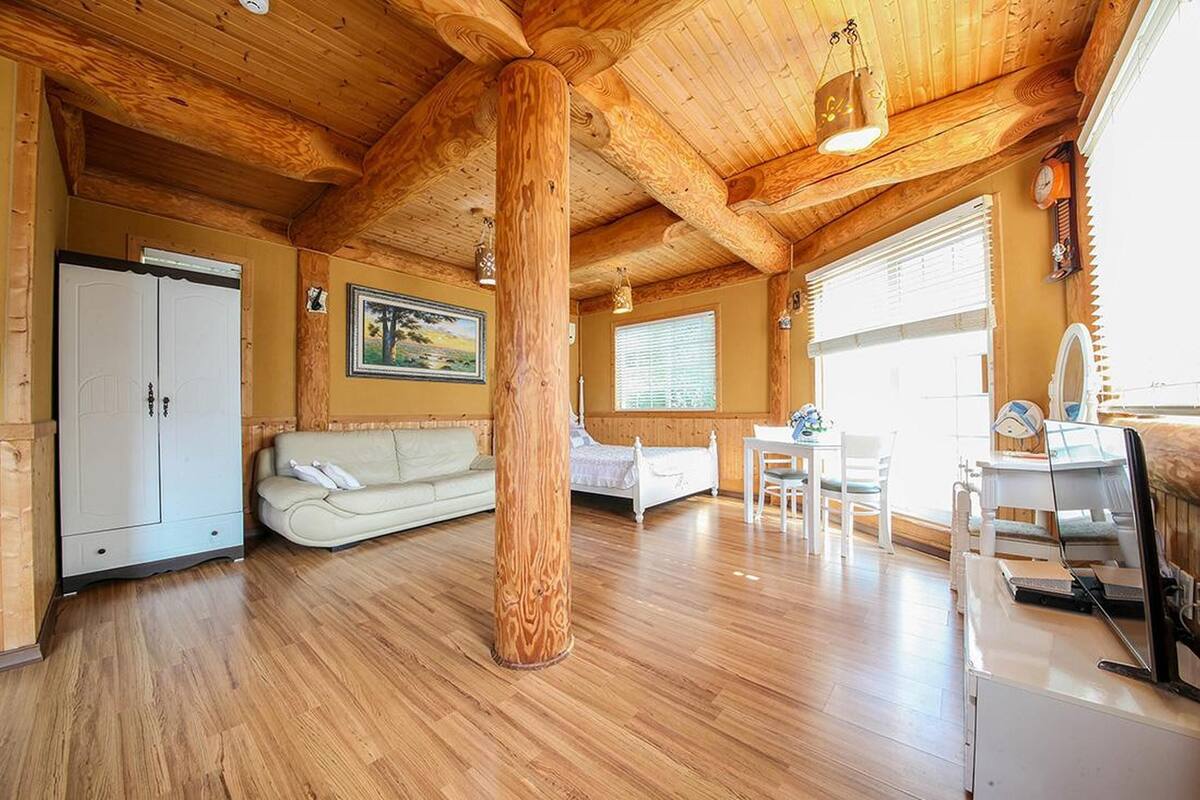 A spacious room with wooden flooring, a large wooden column, a white sofa, a bed, a desk, and a window with blinds.