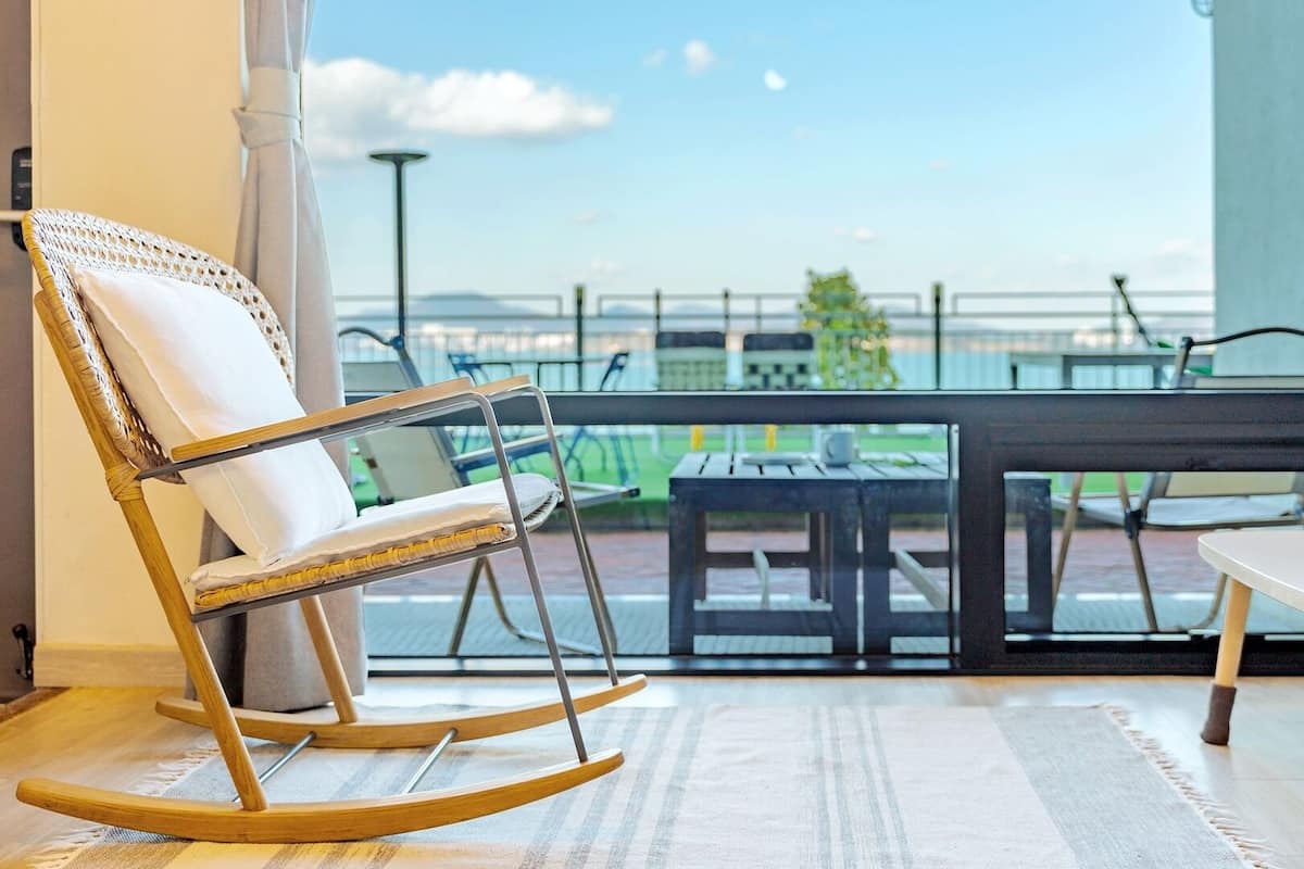 A balcony with a wicker rocking chair, a table, and a view of a sports field.