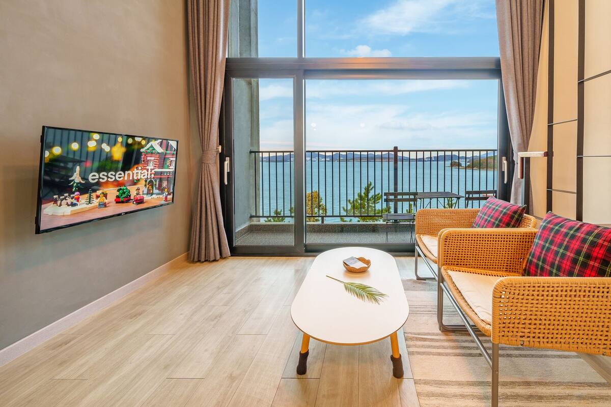 A modern living room with a large window, a flat-screen TV, wicker furniture, and a view of the sea.