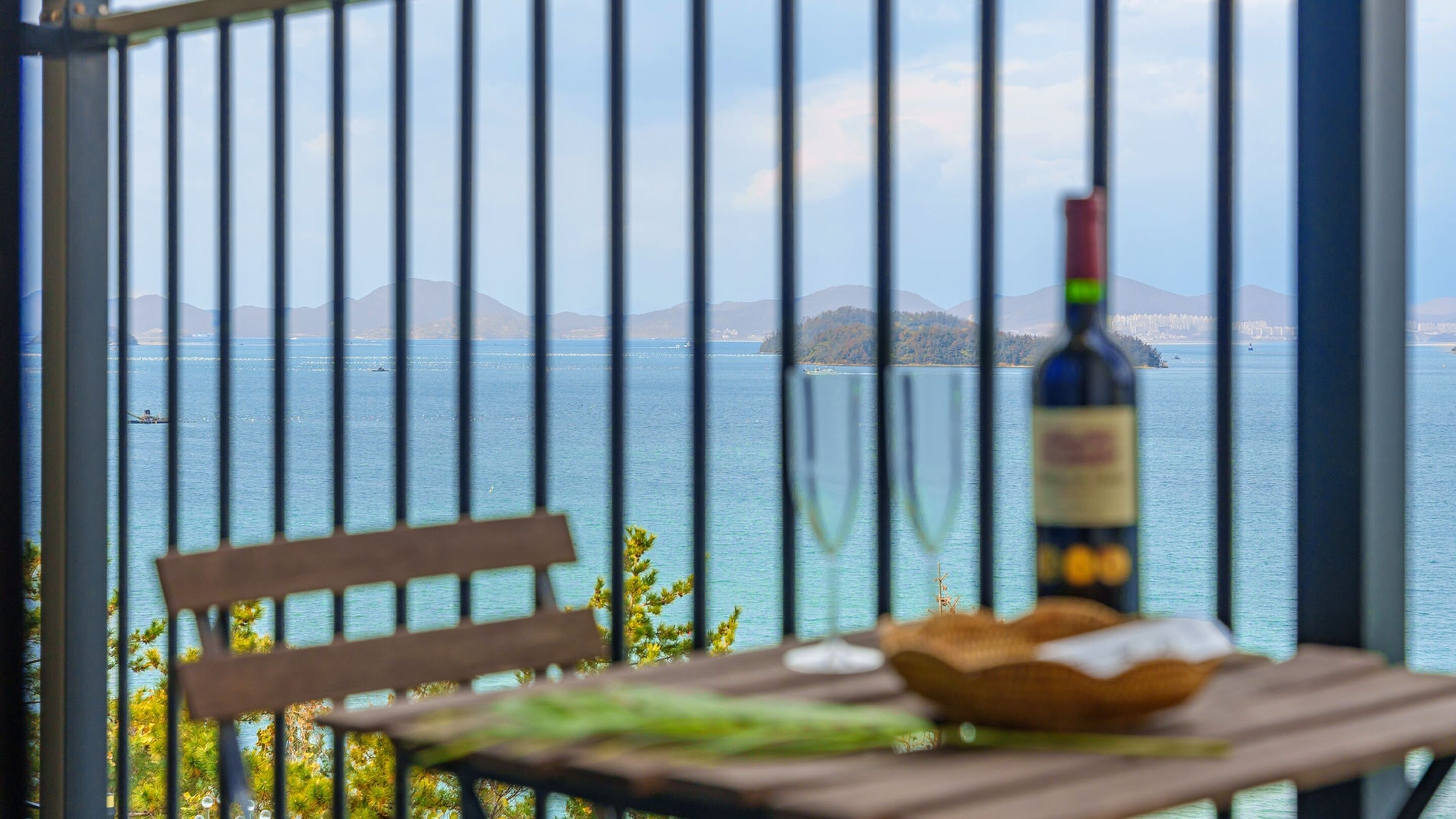 Un balcone con una sedia e un tavolo di legno, una bottiglia di vino e vista su un mare calmo con isole sullo sfondo.