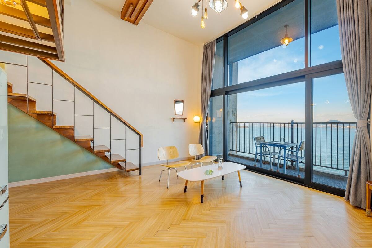 A spacious room with a wooden floor, a staircase, a dining area with a table and chairs, and a large window with a view of the sea.