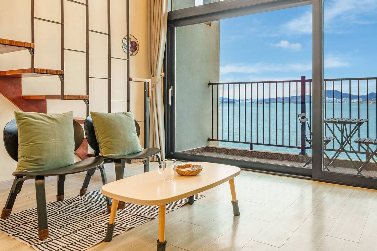 A balcony with a view of the sea, a table with a bowl, two chairs with cushions, and a patterned rug.