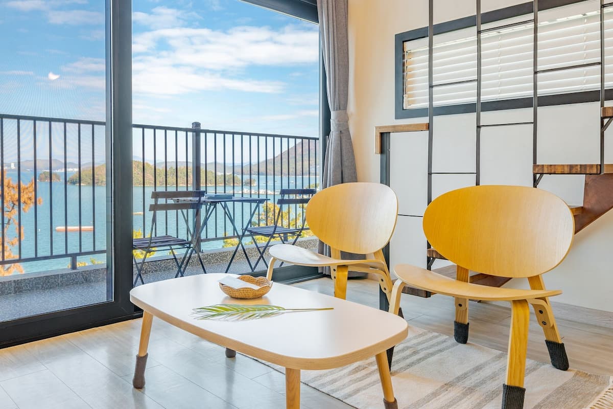 A modern room with a large window overlooking a lake, two wooden chairs, a small table, and a view of the water.
