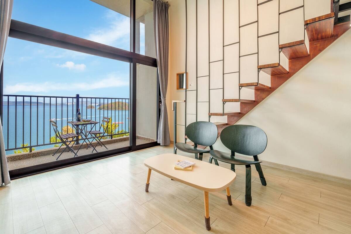 A spacious room with a large window overlooking a body of water, a wooden staircase, and a dining area with chairs and a table.