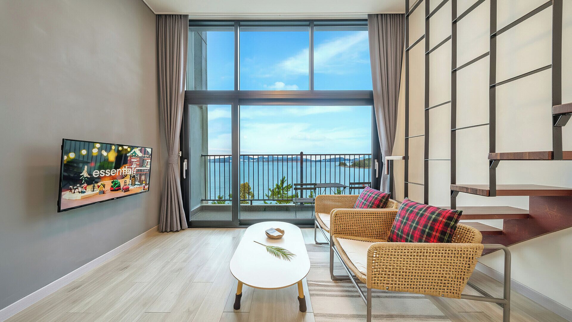 Un salón moderno con un gran ventanal, televisor de pantalla plana, mobiliario de mimbre y una escalera.