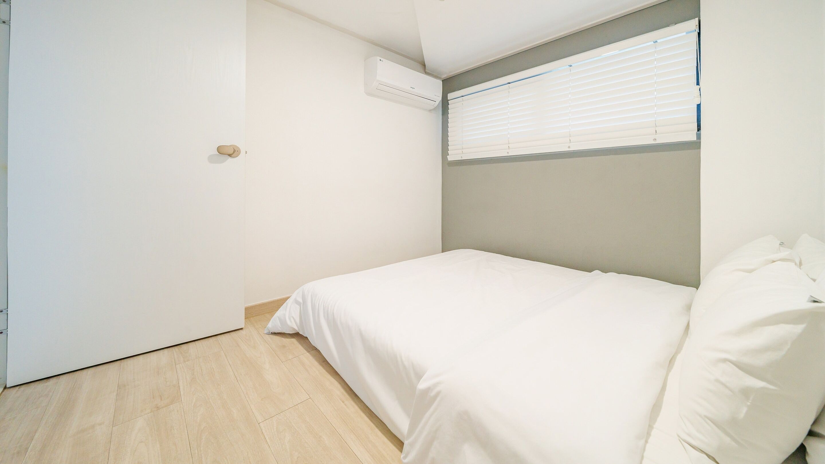 Un dormitorio minimalista con una cama individual, una puerta, una ventana con persianas y un equipo de aire acondicionado.