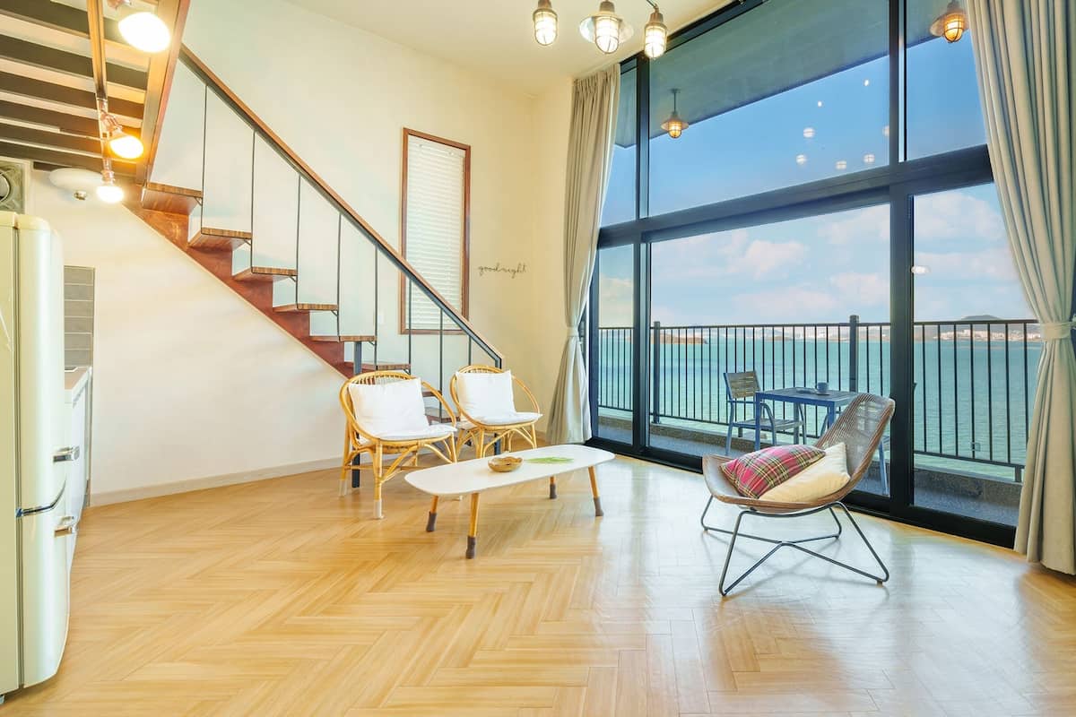 A spacious living area with a wooden herringbone floor, a staircase, wicker furniture, and a large window overlooking a body of water.