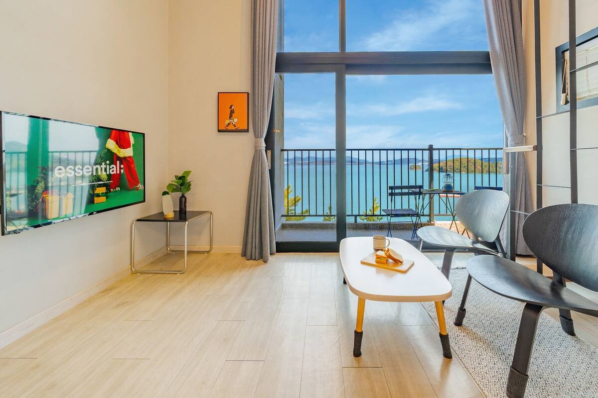 A modern living room with a large window, a flat-screen TV, a small side table, a coffee table, and a chair.