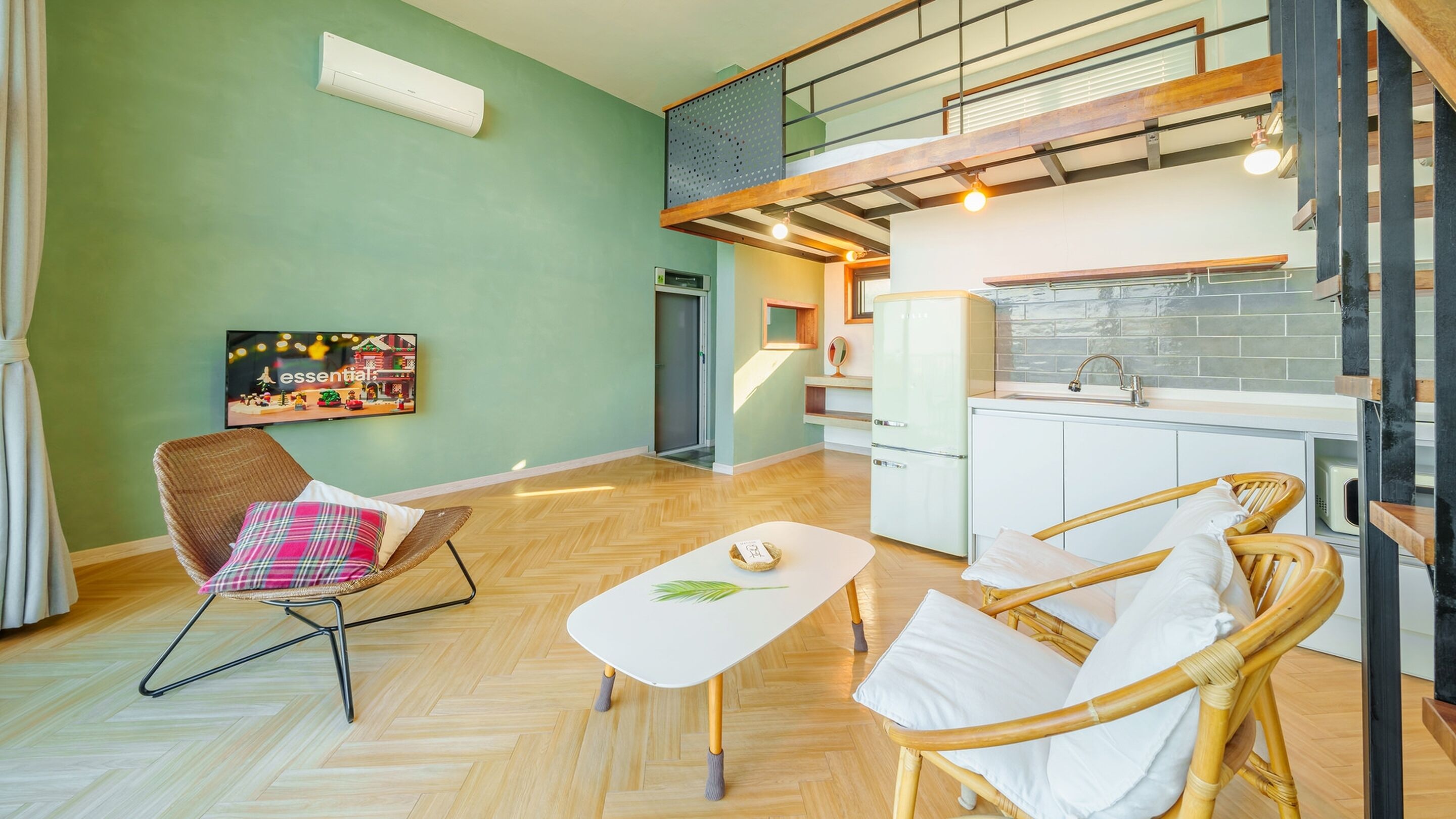 A modern living room with a herringbone floor, wicker chairs, a white coffee table, and a kitchen area with a white refrigerator and a wooden staircase.